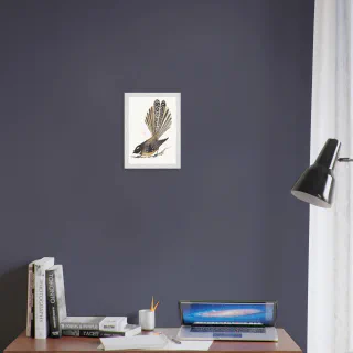 Small white framed dark pīwakawaka print above a desk with books and a laptop