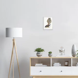 Small white framed dark pīwakawaka print above a sideboard beside a floor lamp